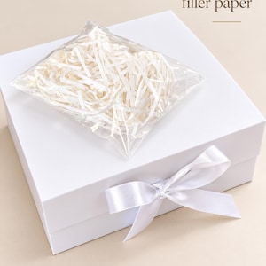 May include: A square white gift box featuring a white satin ribbon tied in a bow on the front. A clear plastic bag containing shredded white filler paper rests on top of the box. The text "Comes with filler paper" is printed above the box.