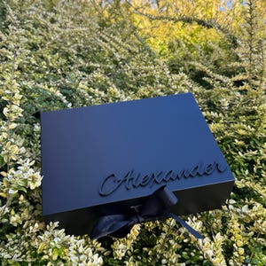 May include: A rectangular matte black gift box featuring the name Alexander in raised black script lettering on the lid, accented with a black ribbon tied in a bow at the front center, creating a sleek and monochromatic appearance.