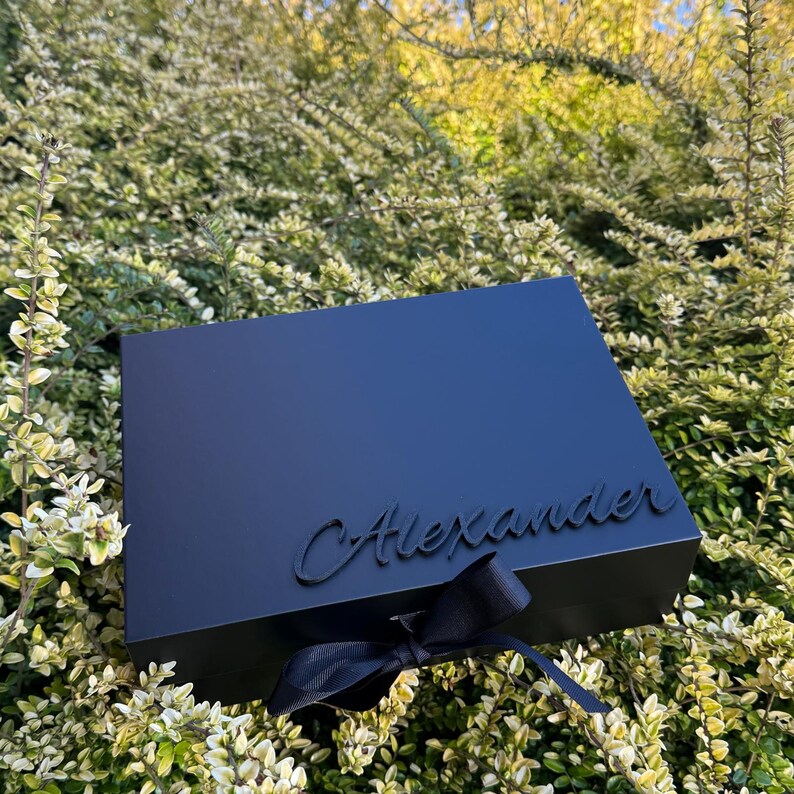 May include: A rectangular matte black gift box featuring the name Alexander in raised black script lettering on the lid, accented with a black ribbon tied in a bow at the front center, creating a sleek and monochromatic appearance.