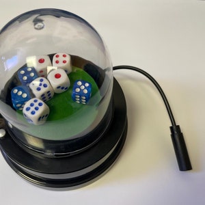 May include: A black electric dice shaker with a clear plastic dome. The shaker contains multiple white dice with blue dots and red dots. The shaker is on a white surface.