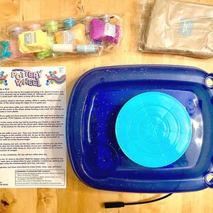 May include: A blue and white pottery wheel with a blue base and a white spinning plate. The wheel is surrounded by a white instruction sheet with the title "Motorized Pottery Wheel" and instructions on how to create a pot. The set includes a bag of clay, tools, and paint.
