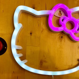 May include: A white light-up outline of Hello Kitty's face with a pink bow. The light is on and the outline is illuminated.