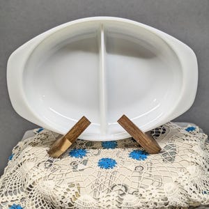 May include: A white, oval divided serving dish with handles. The dish is made of a glossy material and is divided into two sections. Two wooden supports are placed on a cream-colored doily with blue floral accents.