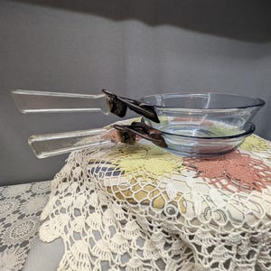 May include: Two clear glass pans with handles, stacked on top of each other. The pans have a round shape and are made of clear glass. The handles are attached with dark metal clips. The pans are sitting on a vintage lace doily.
