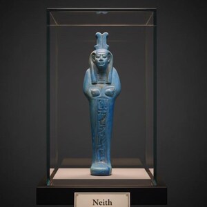May include: A blue statue of Neith, an ancient Egyptian deity, displayed in a glass case. The statue features intricate details, including hieroglyphs and a headdress. The base of the display case has a nameplate that reads "Neith".