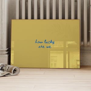 May include: A rectangular mustard yellow wall art piece featuring the dark blue handwritten text how lucky are we centered on the surface. The artwork has a smooth, reflective finish and is positioned horizontally on a light-colored floor.