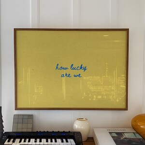 May include: A wooden console table displays a framed yellow artwork with the text "how lucky are we." A small black keyboard, a white vase, a stack of books, and an orange vase are also on the table.