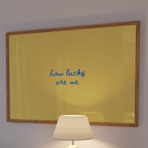 May include: A rectangular wall art piece in a wooden frame featuring a solid mustard yellow background with the blue handwritten text how lucky are we centered in the middle of the print. The artwork is displayed above a white tiled cabinet.
