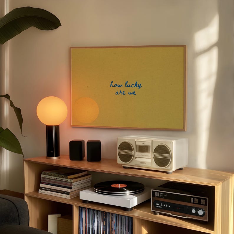 May include: A modern living room setup featuring a wooden shelf with a turntable, stereo, and a vintage boombox. A yellow framed art piece with the text "how lucky are we" hangs above. A black and gold lamp and small speakers are also visible.