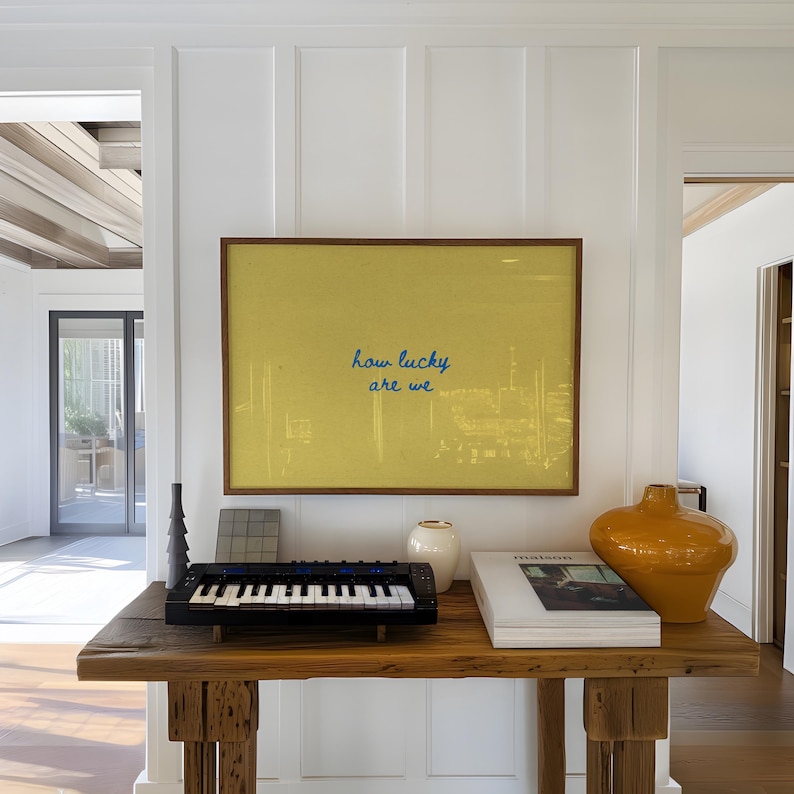 May include: A wooden console table displays a framed yellow artwork with the text "how lucky are we." A small black keyboard, a white vase, a stack of books, and an orange vase are also on the table.