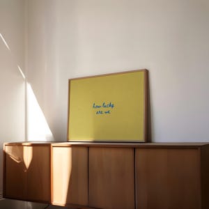May include: A framed yellow print with the text "how lucky are we" in blue script. The print is displayed on a wooden cabinet against a white wall, with sunlight streaming in from a window.