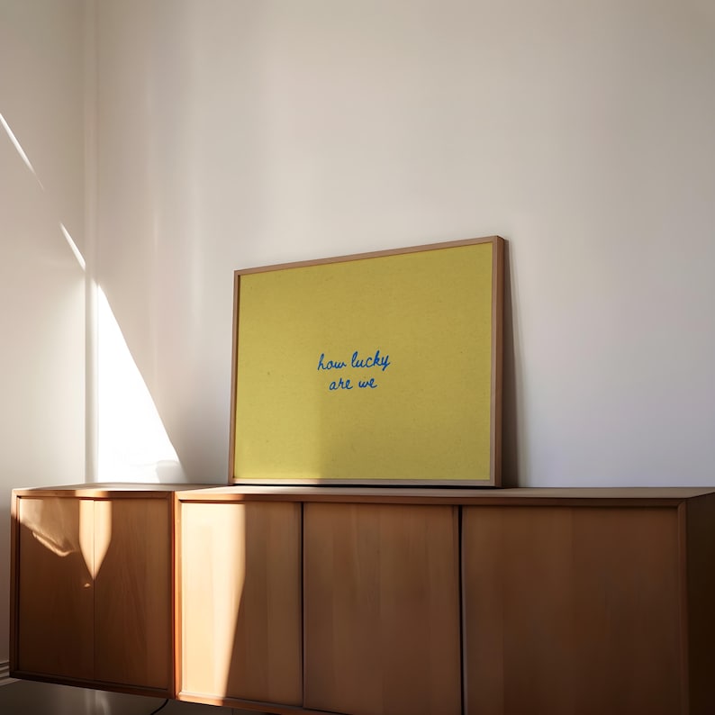 May include: A framed yellow print with the text "how lucky are we" in blue script. The print is displayed on a wooden cabinet against a white wall, with sunlight streaming in from a window.