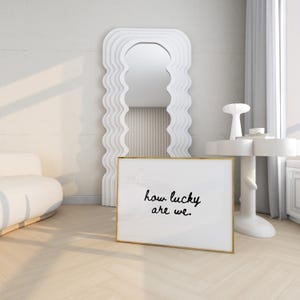 May include: A white framed art print with the text "how lucky are we." is displayed on a light wood floor. A large, wavy-edged white mirror and a small white side table with a decorative lamp are in the background. The room has a minimalist design.