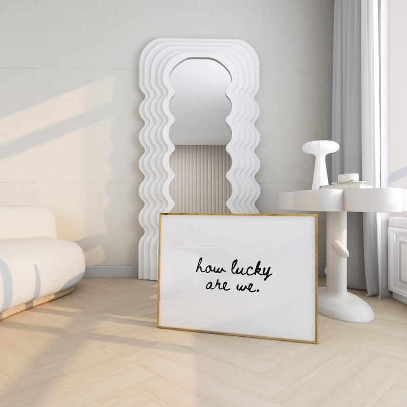 May include: A white framed art print with the text "how lucky are we." is displayed on a light wood floor. A large, wavy-edged white mirror and a small white side table with a decorative lamp are in the background. The room has a minimalist design.