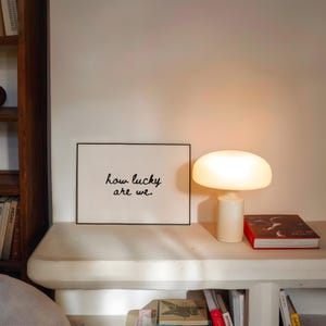 May include: A cream-colored mushroom-shaped table lamp illuminates a white shelf. A framed print with the text "how lucky are we." sits next to the lamp. A red book and other books are also on the shelf.