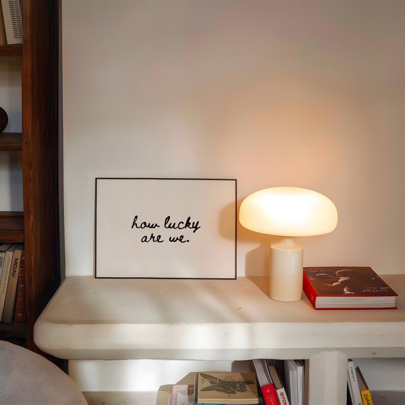 May include: A cream-colored mushroom-shaped table lamp illuminates a white shelf. A framed print with the text "how lucky are we." sits next to the lamp. A red book and other books are also on the shelf.