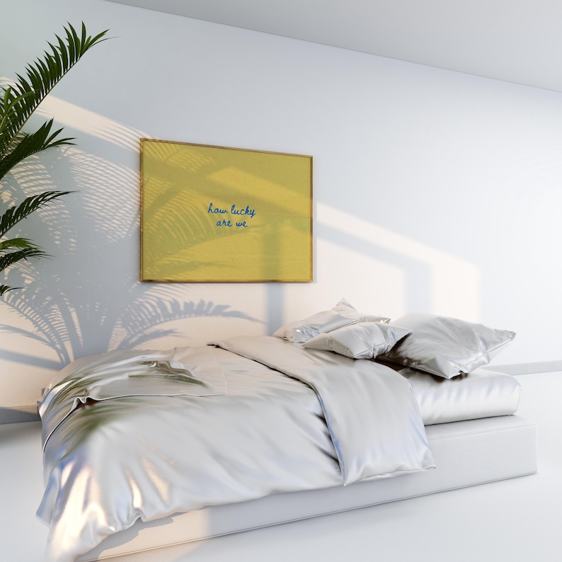 May include: A modern bedroom scene featuring a bed with a silver satin duvet and pillows. A framed artwork on the wall reads "how lucky are we" in blue script. Palm frond shadows add a natural touch.