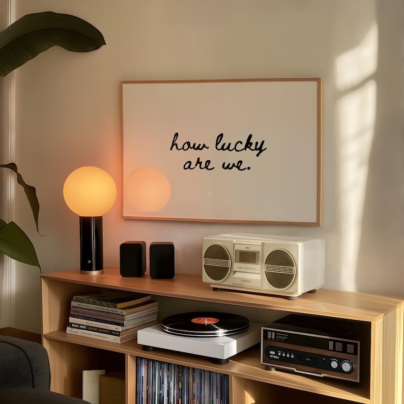 May include: A wooden shelf unit displays a white framed print with the text "how lucky are we." A white record player, a vintage stereo, and a retro boombox are on the shelf. A black and gold lamp and two black speakers are also on the shelf.