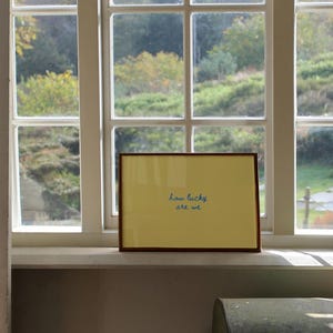 May include: A rectangular framed print featuring a solid yellow background with the text "how lucky are we" written in a blue cursive script. The frame is made of a thin brown material and is positioned horizontally on a white window sill.