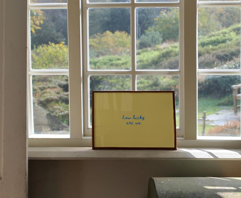 May include: A rectangular framed print featuring a solid yellow background with the text "how lucky are we" written in a blue cursive script. The frame is made of a thin brown material and is positioned horizontally on a white window sill.
