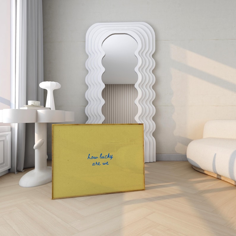 May include: A yellow framed art print with the text "how lucky are we" in blue script. The print is leaning against a white wavy-edged mirror. A white side table and a white sofa are also in the room.
