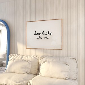 May include: A framed print with the text "how lucky are we" in black script. The print hangs on a white paneled wall above a cream-colored sofa with matching pillows. A blue-framed mirror is visible on the left.