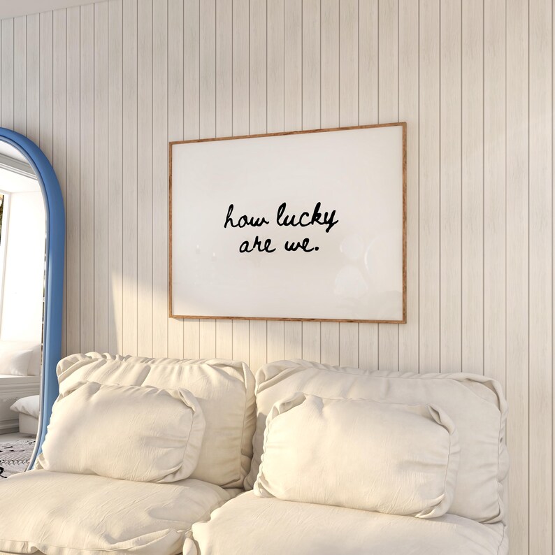 May include: A framed print with the text "how lucky are we" in black script. The print hangs on a white paneled wall above a cream-colored sofa with matching pillows. A blue-framed mirror is visible on the left.
