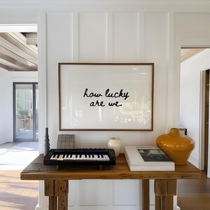 May include: A wooden console table displays a framed print with the text "how lucky are we." A small black keyboard, a white vase, a book, and an orange vase are also on the table. The wall is white.