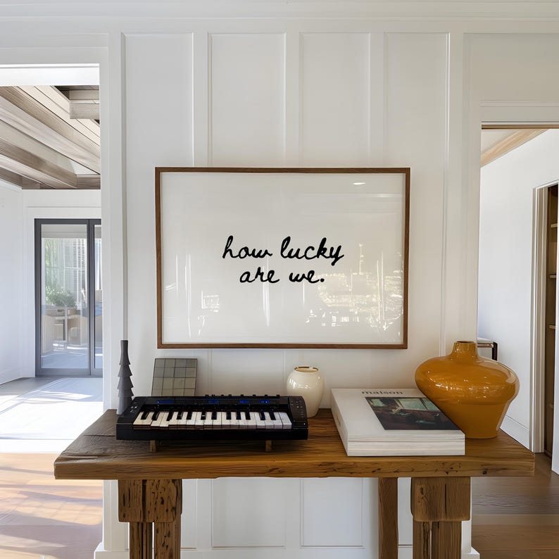 May include: A wooden console table displays a framed print with the text "how lucky are we." A small black keyboard, a white vase, a book, and an orange vase are also on the table. The wall is white.