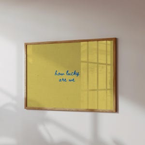 May include: A framed print with a yellow background and the blue text "how lucky are we." The frame is made of light brown wood. The print is hanging on a white wall.