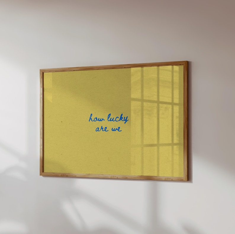 May include: A framed print with a yellow background and the blue text "how lucky are we." The frame is made of light brown wood. The print is hanging on a white wall.