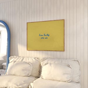 May include: A framed print with a yellow background and the blue text "how lucky are we" hangs on a white paneled wall. Below the print is a white sofa with several white pillows. A blue-framed mirror is visible on the left.