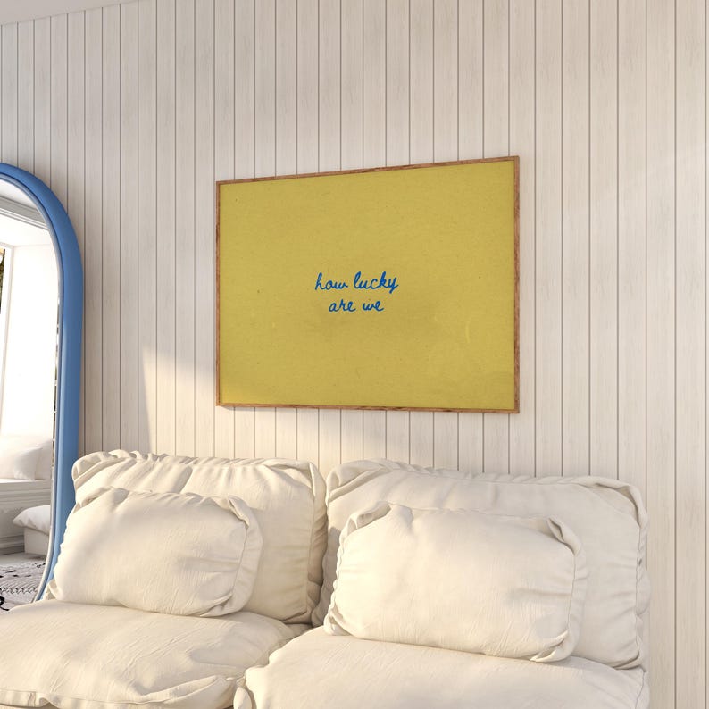 May include: A framed print with a yellow background and the blue text "how lucky are we" hangs on a white paneled wall. Below the print is a white sofa with several white pillows. A blue-framed mirror is visible on the left.