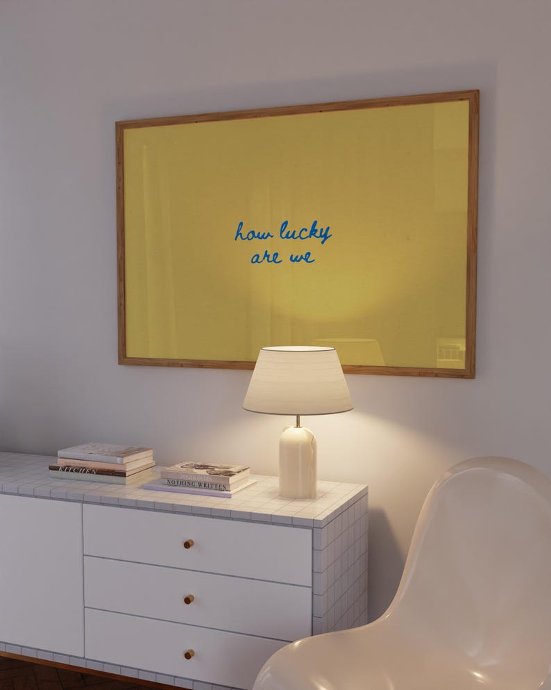 May include: A rectangular wall art piece in a wooden frame featuring a solid mustard yellow background with the blue handwritten text how lucky are we centered in the middle of the print. The artwork is displayed above a white tiled cabinet.