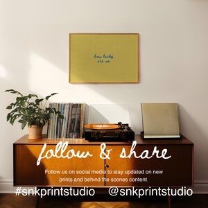 May include: A framed yellow print with the text "how lucky are we" hangs above a mid-century modern cabinet. A record player, vinyl records, and a potted plant are on the cabinet. The words "follow & share" are written on the cabinet.