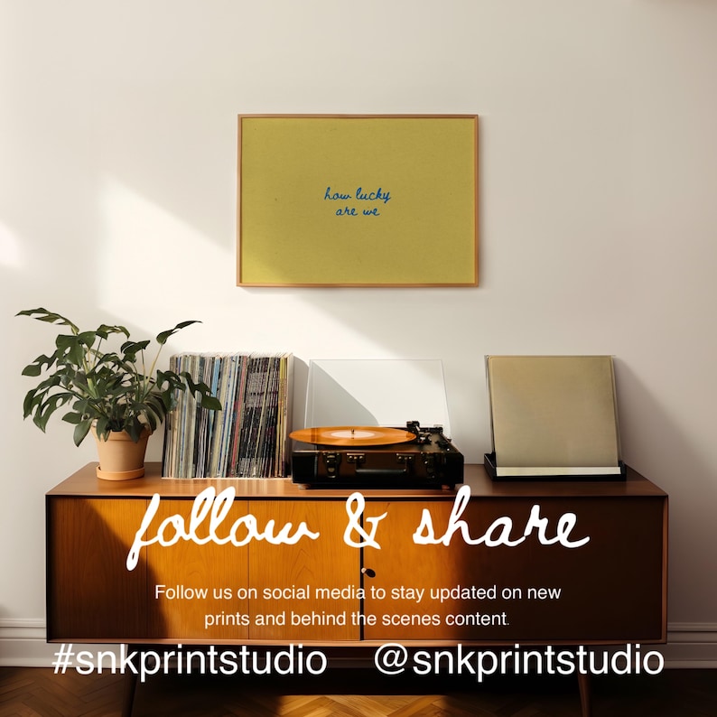 May include: A framed yellow print with the text "how lucky are we" hangs above a mid-century modern cabinet. A record player, vinyl records, and a potted plant are on the cabinet. The words "follow & share" are written on the cabinet.