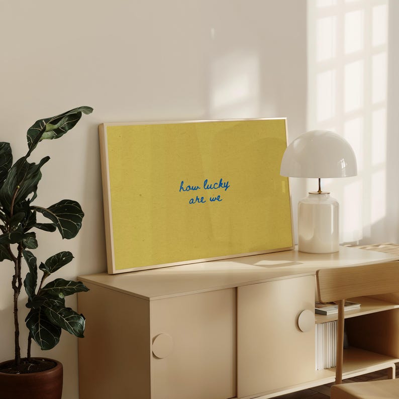 May include: A framed print with a yellow background and the text "how lucky are we" in blue script. The print is displayed on a light-colored cabinet, next to a white lamp with a rounded shade. A potted plant is visible on the left.