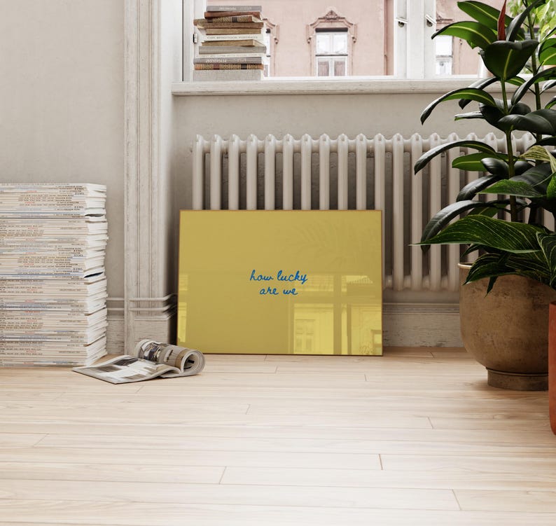 May include: A rectangular mustard yellow wall art piece featuring the dark blue handwritten text how lucky are we centered on the surface. The artwork has a smooth, reflective finish and is positioned horizontally on a light-colored floor.