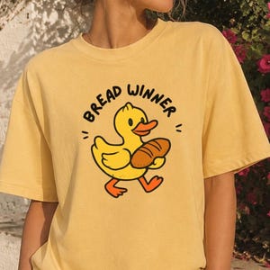 May include: A yellow short-sleeved t-shirt featuring a graphic of a yellow duck holding a brown loaf of bread. The text "BREAD WINNER" is printed in a black arched font above the duck illustration. The shirt is paired with white pants.