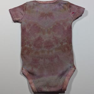 May include: A short-sleeved baby bodysuit in a tie-dye pattern. The bodysuit is primarily a dusty rose color with hints of purple and brown. The fabric appears soft and comfortable, suitable for a baby's delicate skin.