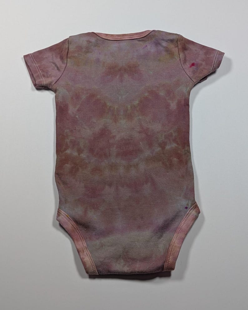 May include: A short-sleeved baby bodysuit in a tie-dye pattern. The bodysuit is primarily a dusty rose color with hints of purple and brown. The fabric appears soft and comfortable, suitable for a baby's delicate skin.