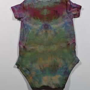 May include: A tie-dye baby bodysuit in shades of green, blue, purple, and brown. The short-sleeved one-piece has a round neckline and is made of soft fabric. The tie-dye pattern is symmetrical, with a central design and a light-colored background.