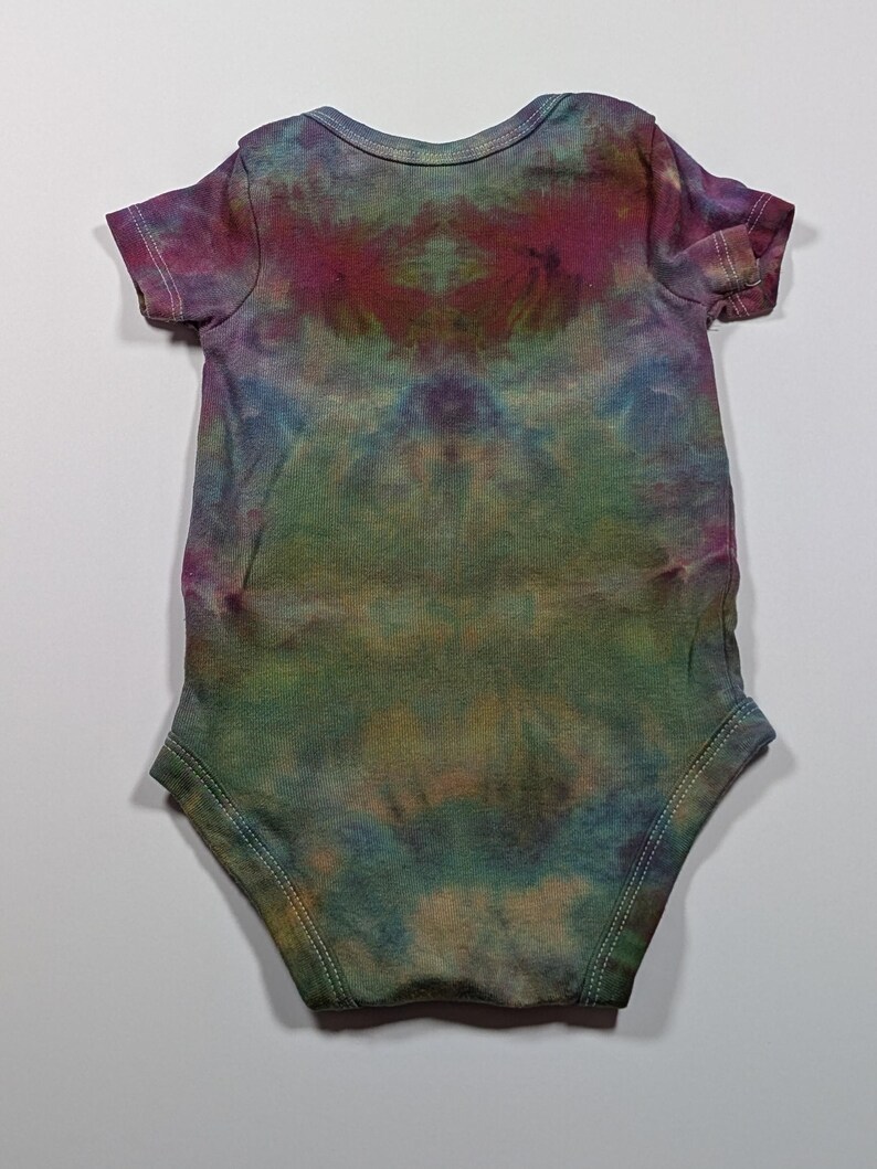 May include: A tie-dye baby bodysuit in shades of green, blue, purple, and brown. The short-sleeved one-piece has a round neckline and is made of soft fabric. The tie-dye pattern is symmetrical, with a central design and a light-colored background.