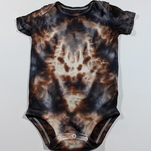 May include: A short-sleeved baby bodysuit with a tie-dye pattern in shades of brown, black, and white. The fabric has a soft, cloud-like appearance. The bodysuit has a round neckline and snaps at the bottom for easy changes.