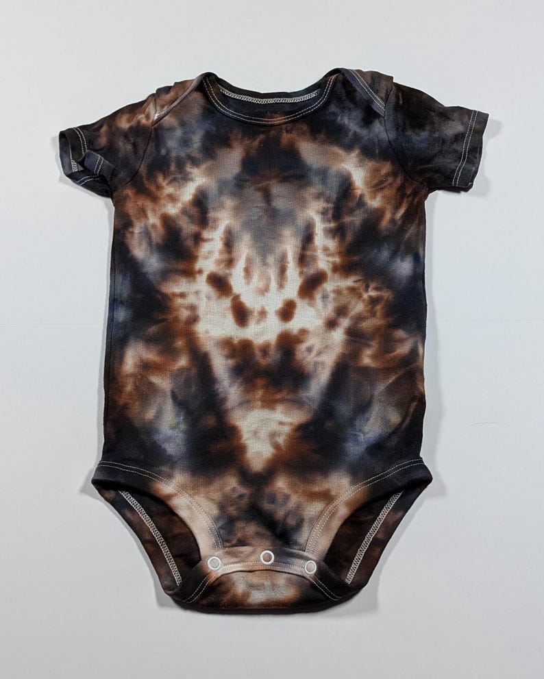 May include: A short-sleeved baby bodysuit with a tie-dye pattern in shades of brown, black, and white. The fabric has a soft, cloud-like appearance. The bodysuit has a round neckline and snaps at the bottom for easy changes.