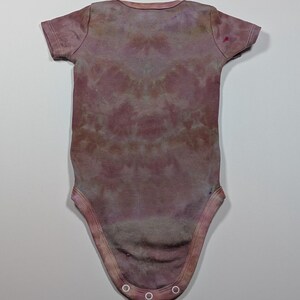 May include: A short-sleeved baby bodysuit in a tie-dye pattern. The bodysuit is primarily a muted mauve color with darker and lighter areas, creating a marbled effect. The garment has a round neckline and snaps at the bottom.