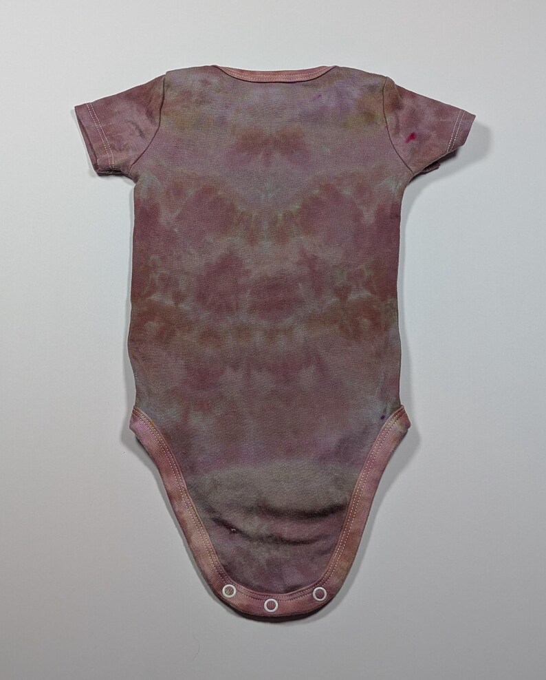 May include: A short-sleeved baby bodysuit in a tie-dye pattern. The bodysuit is primarily a muted mauve color with darker and lighter areas, creating a marbled effect. The garment has a round neckline and snaps at the bottom.