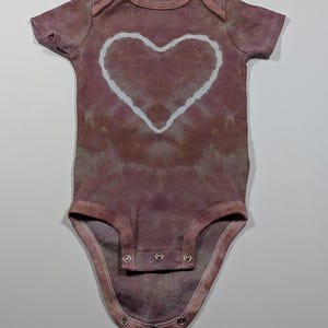 May include: A short-sleeved baby onesie in a tie-dye style, featuring a large white heart in the center. The onesie is primarily a muted mauve color with a pattern of darker and lighter shades. It has snap closures at the bottom.