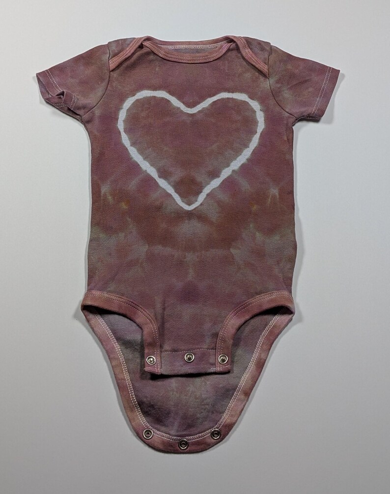May include: A short-sleeved baby onesie in a tie-dye style, featuring a large white heart in the center. The onesie is primarily a muted mauve color with a pattern of darker and lighter shades. It has snap closures at the bottom.
