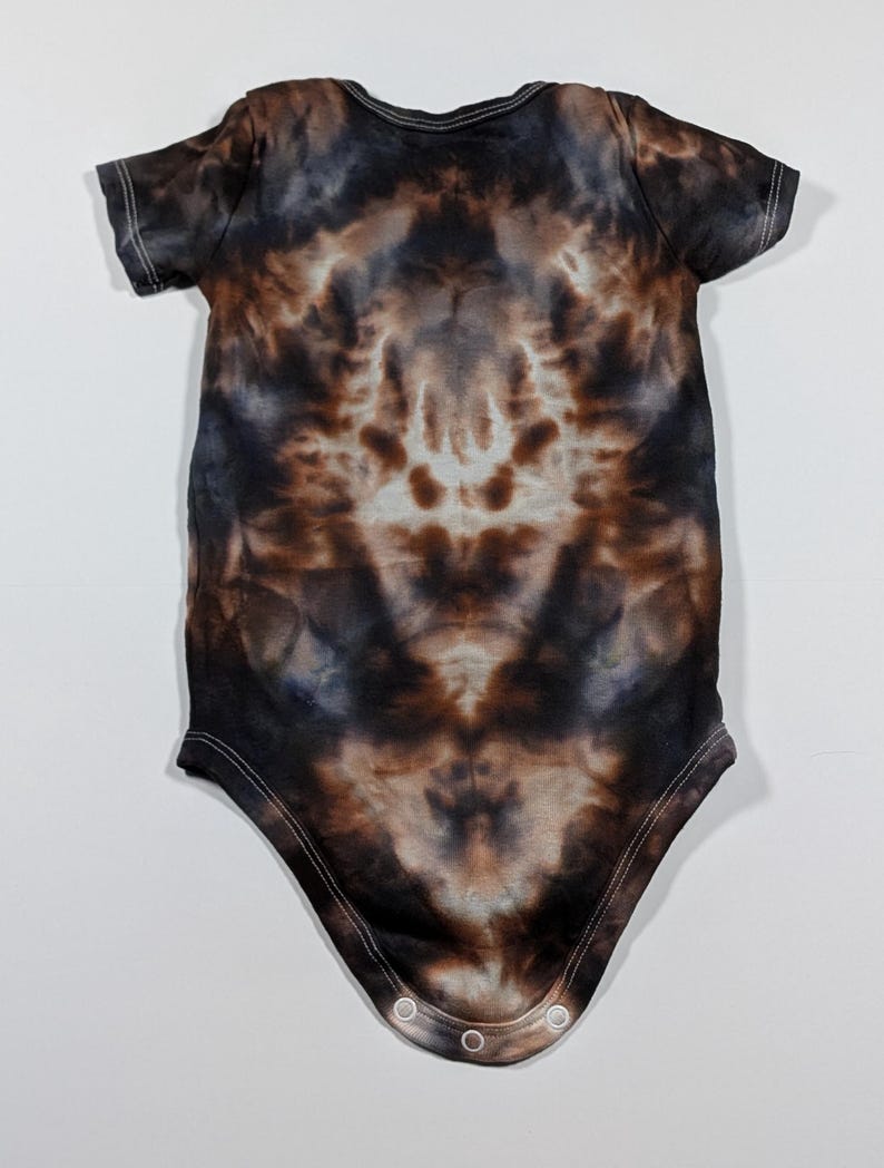 May include: A short-sleeved baby bodysuit with a tie-dye pattern in shades of brown, black, and blue. The bodysuit has a round neckline and snap closures at the bottom. The fabric appears soft and comfortable.